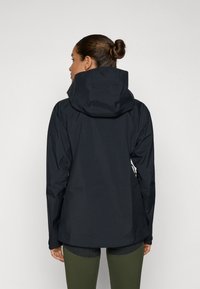 Zwarte waterdichte jas met een capuchon, met een getailleerd ontwerp en een gladde textuur. Gekoord met olijfgroene leggings, gezien van achteren.