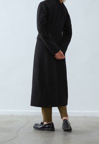 Lange zwarte jas van gestructureerde stof, met een rechte snit. Draagt over bruine, taps toelopende broek en zwarte schoenen met lage hak.