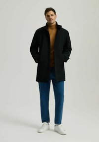 Mężczyzna w czarnym płaszczu, brązowym golfie, niebieskich dżinsach i białych trampkach, stojący z rękami w kieszeniach płaszcza na jednolitym tle.