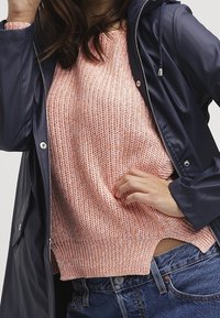 Kvinna som bär en marinblå regnrock över en rosa stickad tröja och blåjeans, med handen vilande på höfterna.