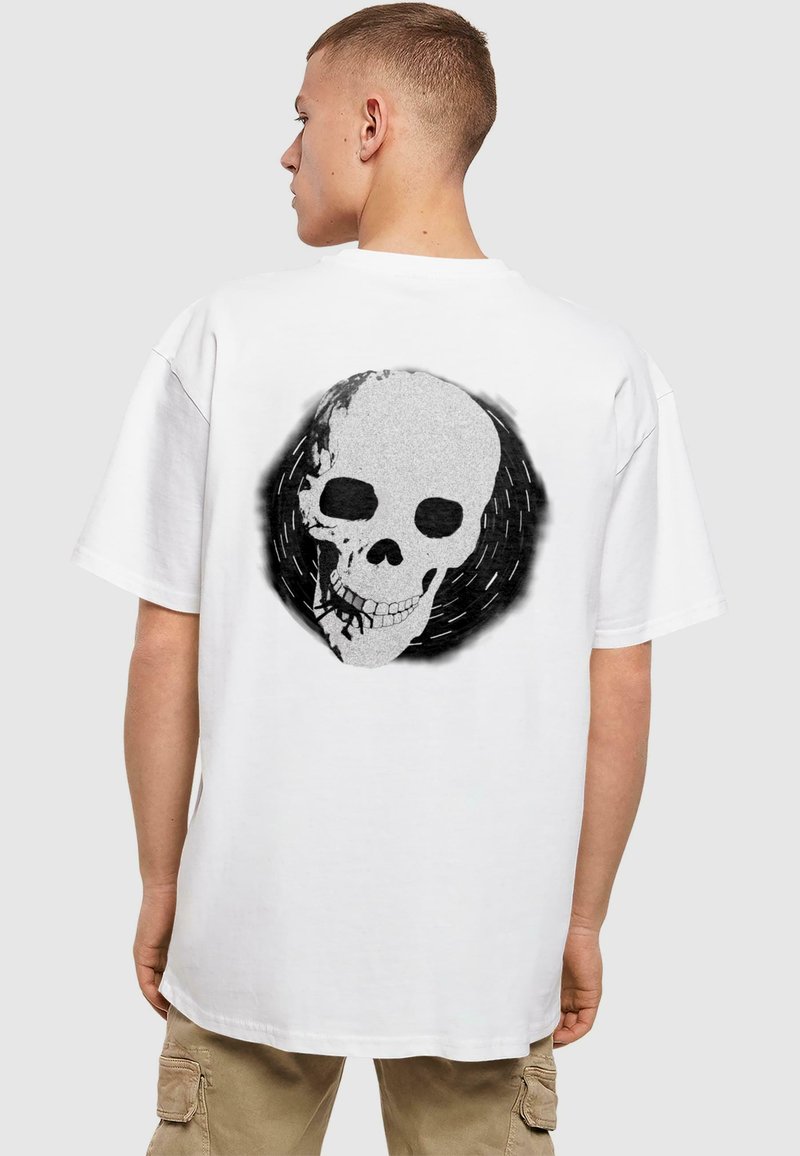 Hvid t-shirt med et stort sort og gråt kraniumprint på ryggen, sat mod et struktureret cirkulært design. Løs pasform, korte ærmer.