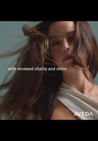 Cheveux bruns longs et flottants en mouvement sur un fond bleu atténué. Le mannequin porte un vêtement drapé blanc. Le texte indique : « avec une vitalité et un éclat renouvelés. »