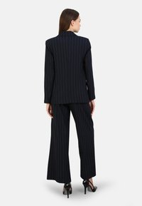 Blazer bleu marine à rayures et pantalon à large jambe, coupe ajustée, tissu texturé, col à revers et chaussures à talons noirs.