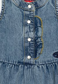 Denimkjole med rund hals, perlemorsknapper, volangdetalj på siden og en samlet skjørt. Har gule sting og en logo-patch.