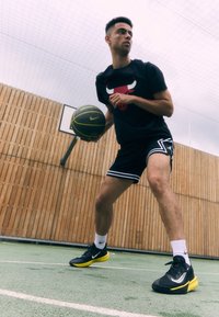 Black Nike basketball shirt with red bull logo, black shorts with white stripes, and black/yellow shoes, dribbling a green basketball outdoors.