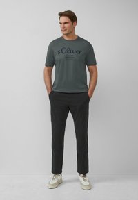 Ciemnozielona bawełniana koszulka z logiem "s.Oliver" w granatowym kolorze, zestawiona z czarnymi spodniami i beżowymi trampkami z białymi akcentami.