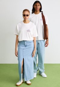 Camiseta blanca con logo, falda maxi de mezclilla azul con abertura lateral y jeans baggy azules claros. Ambos llevan zapatillas blancas. Fondo verde.