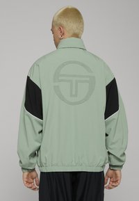 Lys grønn vindjakke med svarte aksentpaneler, med en prikkete sirkel-logo på baksiden og elastiske mansjetter og kant.