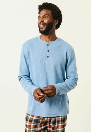 Mann mit Bart und lockigem Haar, der ein hellblaues langärmeliges Henley-Shirt und karierte Hosen trägt, schaut zur Seite und lächelt.