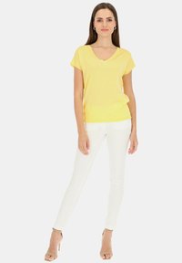 Żółta bluzka z krótkim rękawem i dekoltem w kształcie litery V, wykonana z lekkiego materiału, połączona z dopasowanymi białymi spodniami. Całość dopełniają cieliste sandały na wysokim obcasie.