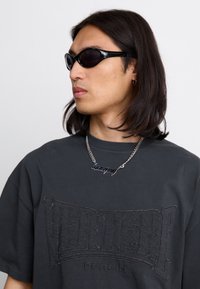 Black oversized t-shirt with embossed logo "VERTER" and "BERLIN" text, paired with black angular sunglasses and a silver chain necklace.
