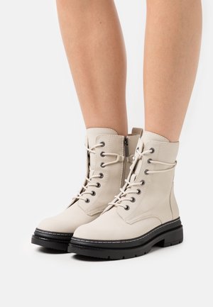 Beige Schnürstiefeletten aus glattem Leder, mit schwarzen Sohlen, seitlichen Reißverschlüssen und dekorativen Nähten entlang der Kanten.