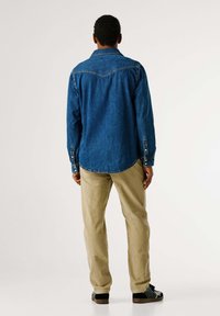 Traper košulja tamnoplave boje s gumbima na manžetnama i šiljastim ovratnikom. Uparena sa svijetlobéž hlačama od manšestra i crnim cipelama. Pogled sa stražnje strane.