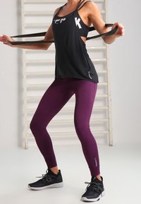 Zwarte mouwloze sporttop met uitsneden aan de zijkanten, gecombineerd met strakke bordeauxrode leggings en zwarte sneakers. De top heeft een logo ontwerp.