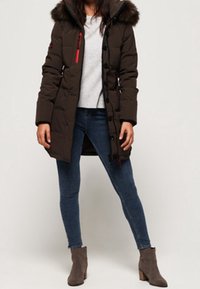 Brun polstret parka med en pelsforet hætte, røde detaljer, knaplukning og sidelommer, kombineret med blå jeans og grå ankelstøvler.