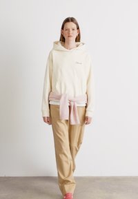 Haut à capuche couleur crème avec une poche avant, associé à un pantalon beige ample. Un pull rose clair noué autour de la taille ajoute une touche de détail.