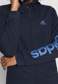 Persona vistiendo una sudadera con capucha azul marino de Adidas con cremallera, con el logo azul en el pecho y el texto "adidas" en la manga, brazos cruzados y mano cerca de la cara.