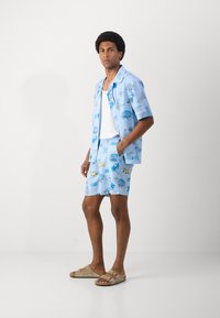 Chemise à manches courtes bleu clair assortie et shorts avec des graphismes de palmiers, de voitures et de texte. Modèle portant des sandales beiges.
