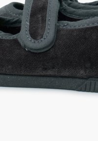 Zapato de ante negro con una correa negra acolchada, suela de goma plana y costuras decorativas a lo largo de los bordes. Diseño simple y funcional.