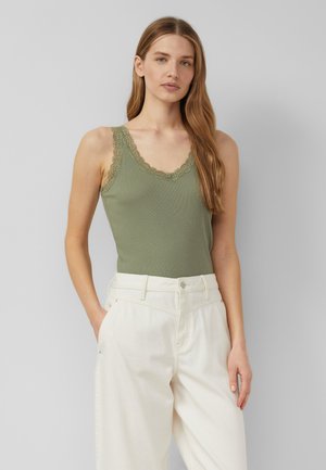 Vrouw die een olijfgroene tanktop met kanten rand draagt, ingestopt in een hoog opgesneden gebroken witte broek, staand met één hand in de zak.