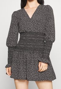 Černé šaty s bílým květinovým vzorem, s dlouhými nabíranými rukávy, výstřihem do V, smockovaným pasem a volným volánkovým lemem. Měkká textilie.