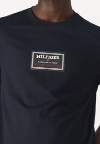 Camiseta de algodón azul marino con un parche rectangular de logo que presenta "HILFIGER" y "AMERICAN CLASSIC" en texto blanco y rojo. Diseño clásico de cuello redondo.