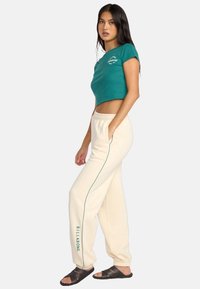 Tielowa, krótka koszulka z logo, kremowe spodnie dresowe z bocznymi lampasami i logo, zestawione z czarnymi sandałami na rzemyki. Gładki materiał, luźny krój.