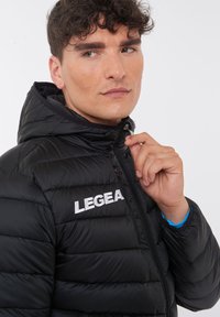 Svart puffjacka med huva, quiltad design och framfickzipper. "LEGEA"-logotyp i vitt på vänster bröstområde.