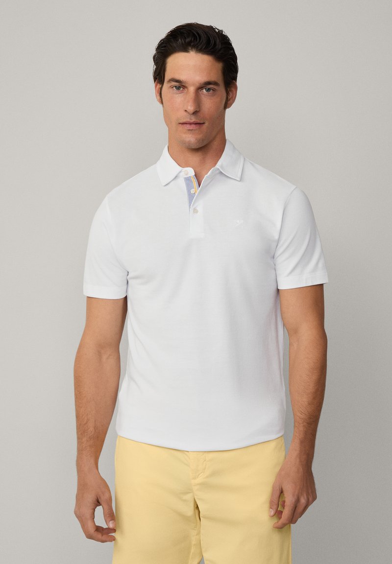 White polo shirt with a short collar, featuring a striped pattern on the inner collar. Made of a smooth fabric, paired with light yellow shorts.