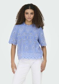 Camicia ricamata azzurro chiaro con bordi scallop, caratterizzata da intricate varianti e occhielli, abbinata a pantaloni bianchi.