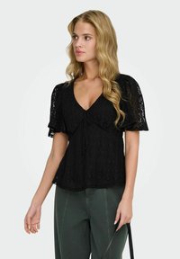 Femme aux longs cheveux blonds porte un haut noir en dentelle à manches courtes avec un col en V et un pantalon vert foncé, tenant une sangle noire à la main.