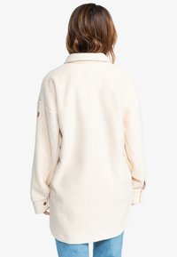 Lys beige fleecejakke med avslappet passform, oversized krage og mansjetter med knapper. Myk tekstur og minimale designelementer på ermene.