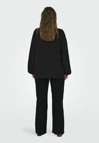 Top negro de gran tamaño con mangas largas y un acabado texturizado, combinado con pantalones de campana negros. Vista desde atrás, la modelo tiene el cabello ondulado.