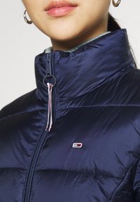 Giacca imbottita blu navy con collo alto, dotata di cerniera frontale, coulisse elastica e una toppa con logo sul petto. Tessuto liscio e brillante.