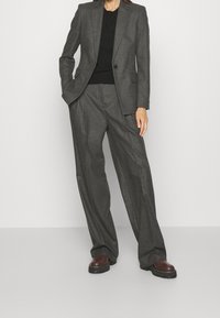 Szary garnitur na miarę z szerokimi spodniami o prostych nogawkach i dopasowaną czarną bluzką. Garnitur ma pojedyncze zapięcie na guzik i klapy z wycięciami.