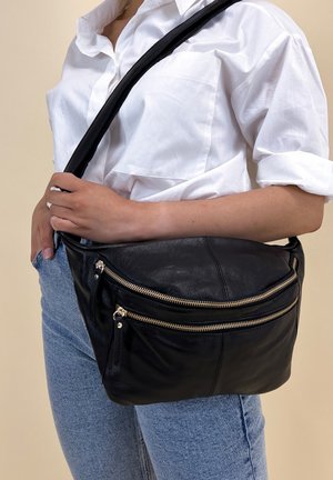Personne portant une chemise blanche retroussée et un jean bleu, avec un sac en cuir noir porté en bandoulière, orné de deux poches zippées dorées, porté sur la poitrine et la hanche.