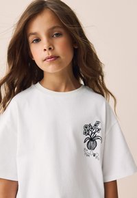 Camiseta blanca de algodón con un jarrón negro bordado con flores y el texto "un AMOUR" en una fuente divertida en el lado izquierdo.