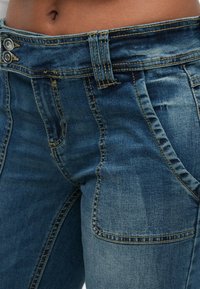 Sötétkék farmernadrág klasszikus dizájnnal, kontrasztos varrással, dupla gombos záródással és elülső zsebekkel.