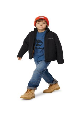 Veste noire, t-shirt bleu Timberland avec logo, jeans en denim avec des patchs et bottes beiges. Un bonnet orange complète la tenue.