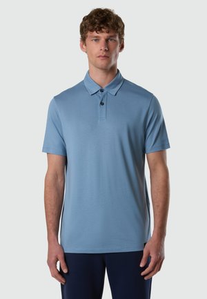 Hombre con cabello rizado que lleva una camiseta tipo polo de manga corta azul claro y pantalones oscuros, de pie frente a un fondo claro y liso.