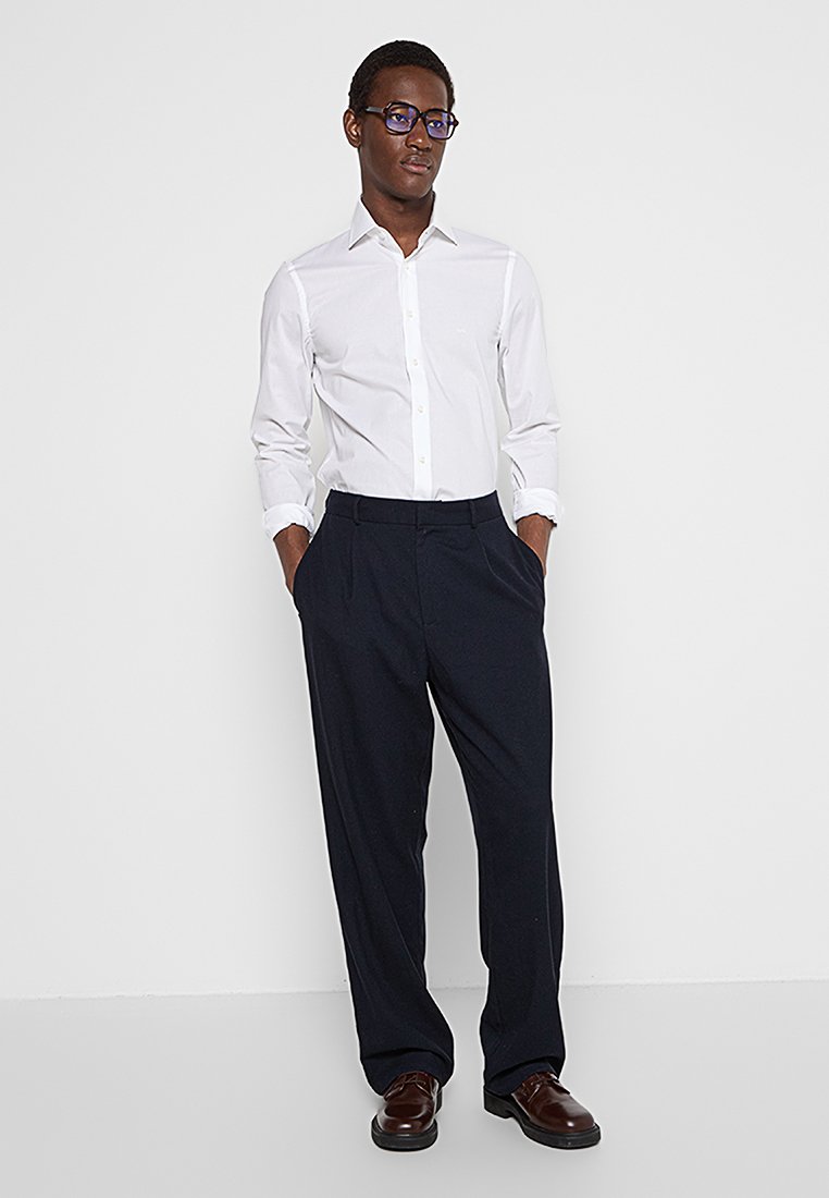 Camisa blanca de botones con mangas largas combinada con pantalones oscuros de color azul marino. La camisa cuenta con un cuello clásico y un detalle minimalista.