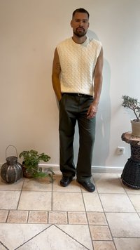 Kabelstrick ärmelloser Pullover in Creme, olivgrüne Hose und schwarze Schuhe. Der Hintergrund zeigt Pflanzen und dekorative Gegenstände auf einem gefliesten Boden.