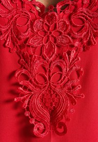 Röd tyg med intrikata, upphöjda broderier som visar blommor och snurrande mönster. Texturerade detaljer ökar den visuella djupet och detaljrikedomen.