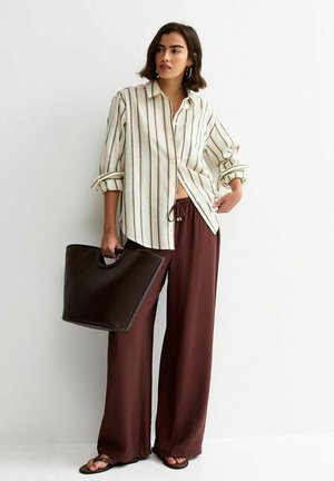 Vrouw draagt een opgerolde gestreepte blouse en losse bruine broek, houdt een grote donkerbruine handtas vast en staat tegen een witte muur.