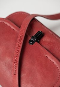 Rode leren schoudertas met een gladde textuur, voorzien van een zwart hardware sluiting en reliëfbranding op de schouderband. Subtiele stikseldetails.
