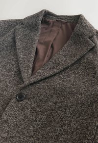 Pelēks teksturēts mētelis ar izgrieztu apkakli, vienu melnu pogu un brūnu odere. Audumam ir raksts ar austiem elementiem.