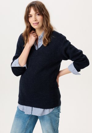 Vrouw draagt een marineblauwe trui over een gestreept shirt en blauwe spijkerbroek, staat met één hand op de heup en de andere bij haar nek, glimlachend.