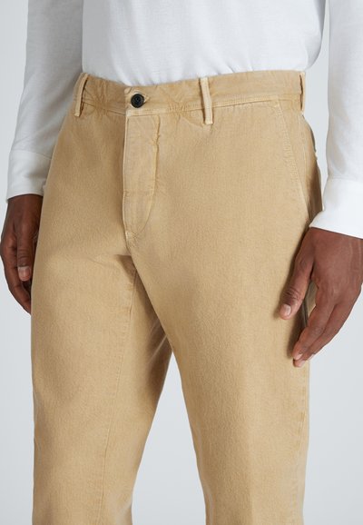 Hombre que lleva pantalones beige con un botón negro y una camisa blanca de manga larga, con las manos descansando a los lados contra un fondo liso.