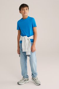 Modra majica s kratkimi rokavi, svetlo modri jeans, sivo trenirko zavezano okoli pasu in sivi športni copati z črnimi detajli.