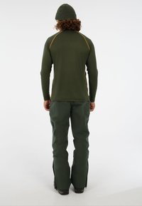 Tmavo zelená košeľa s dlhým rukávom a žltými bočnými švami, doplnená o voľné zelené nohavice so zipsami pri členkoch.